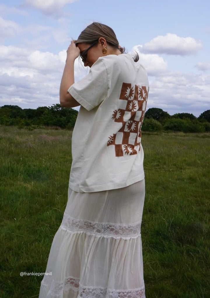 Person in meadow wearing The Artist Series TBCo x Frankie Penwill Cotton T-Shirt with brown checkerboard graphic, paired with a tiered white skirt.