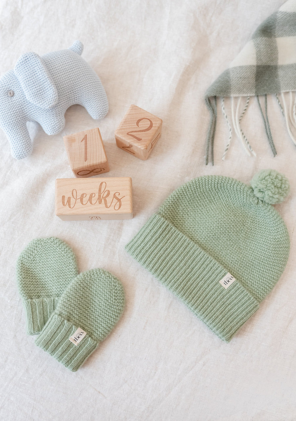 TBCo Merino Wool Baby Beanie in pistachio green with matching mittens, displayed with wooden blocks and plush elephant toy