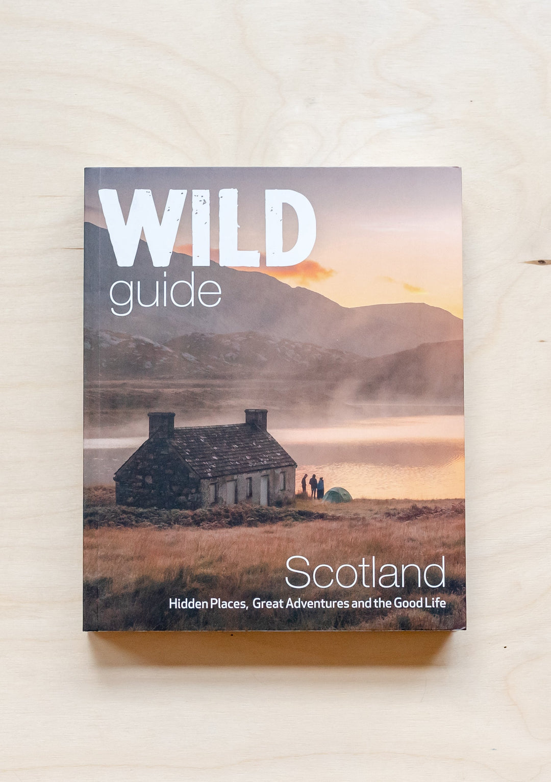 TBCo Wild Guide Scotland book cover showing misty loch with stone cottage at sunset in Scottish Highlands