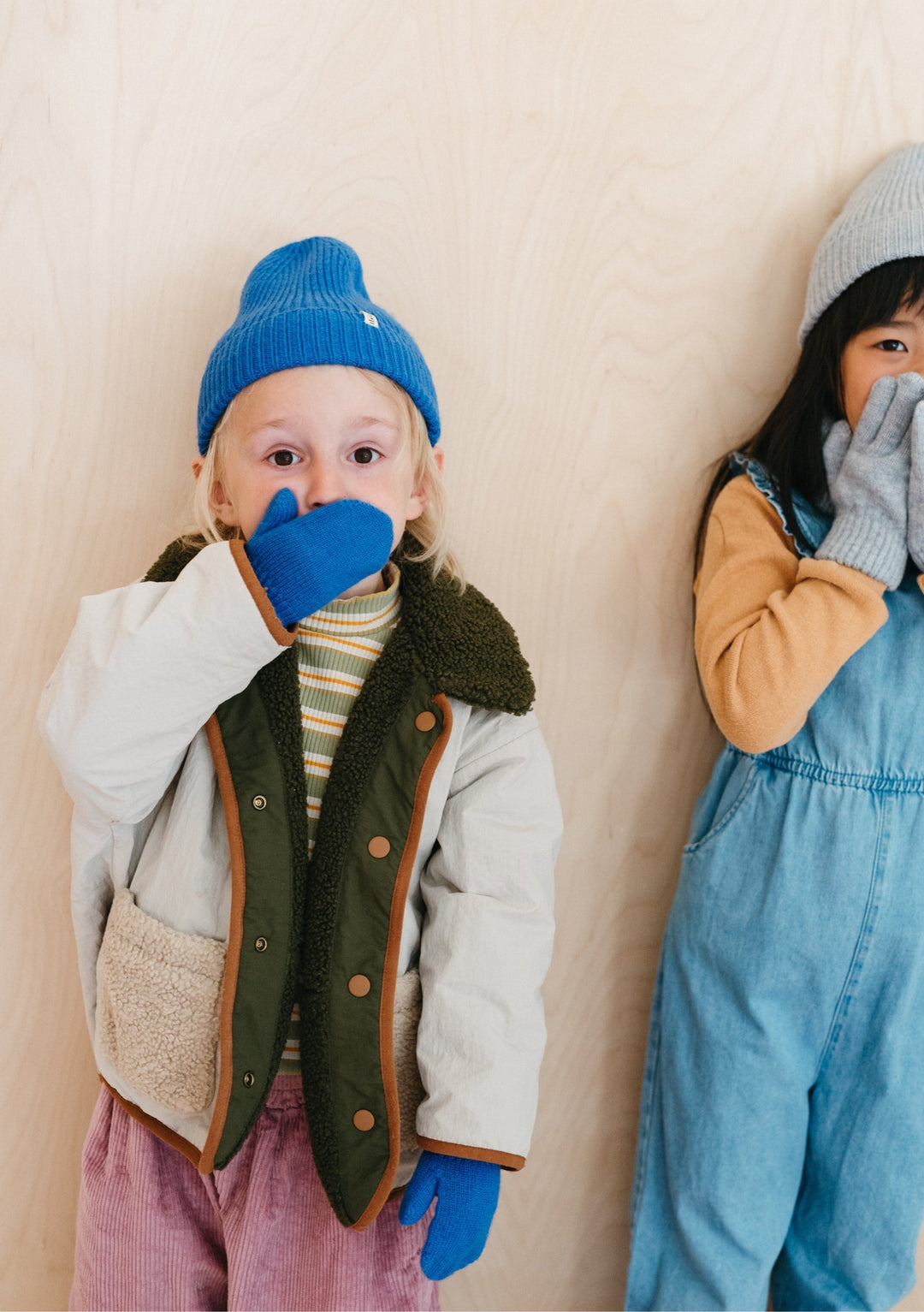 Small child wearing TBCo Merino Wool Kids Mittens in Blue with matching blue beanie, layered winter clothing against beige wall