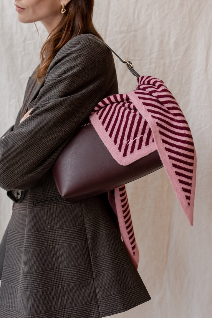 Pink and burgundy striped merino wool triangle scarf by TBCo, styled over grey blazer with leather handbag, showing versatile wear