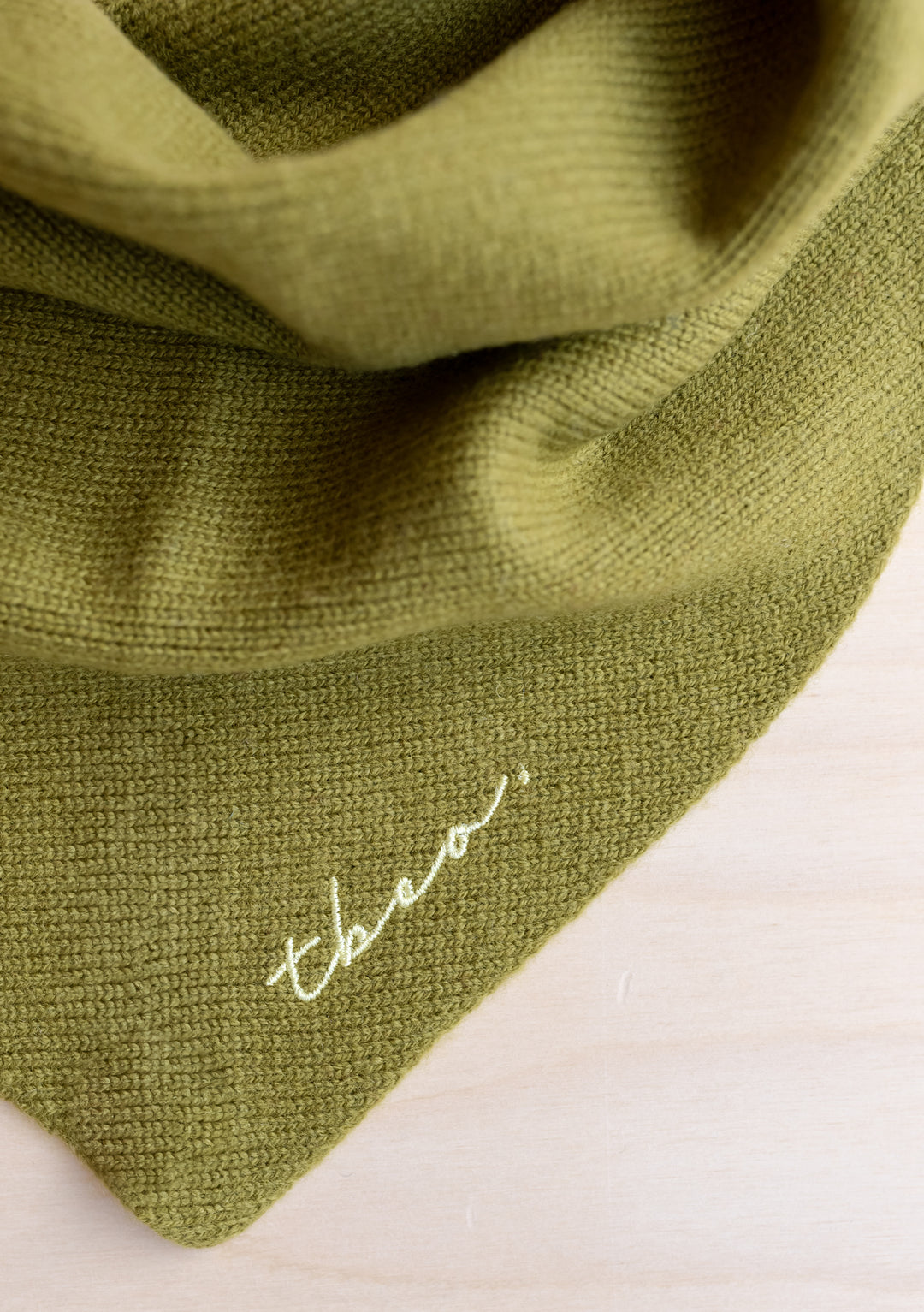 Close-up detail of TBCo Merino Triangle Scarf in Olive showing soft knit texture and signature embroidered logo in lime yellow