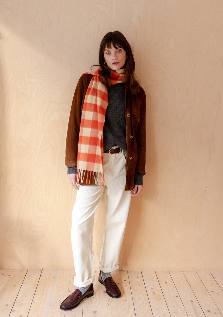 TBCo lambswool scarf in orange and cream gingham pattern styled with brown jacket and white trousers on wooden backdrop