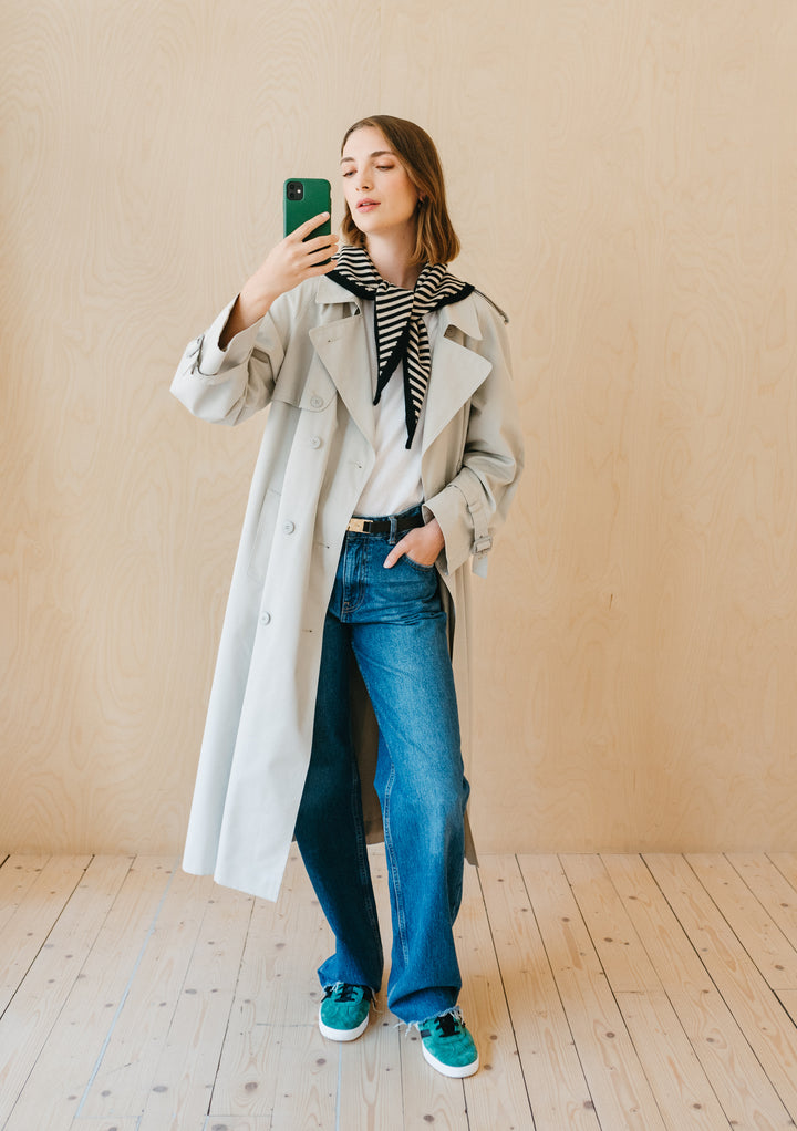 A person wearing TBCo Merino Triangle Scarf in Black Stripe styled with beige trench coat and casual denim, taking mirror selfie