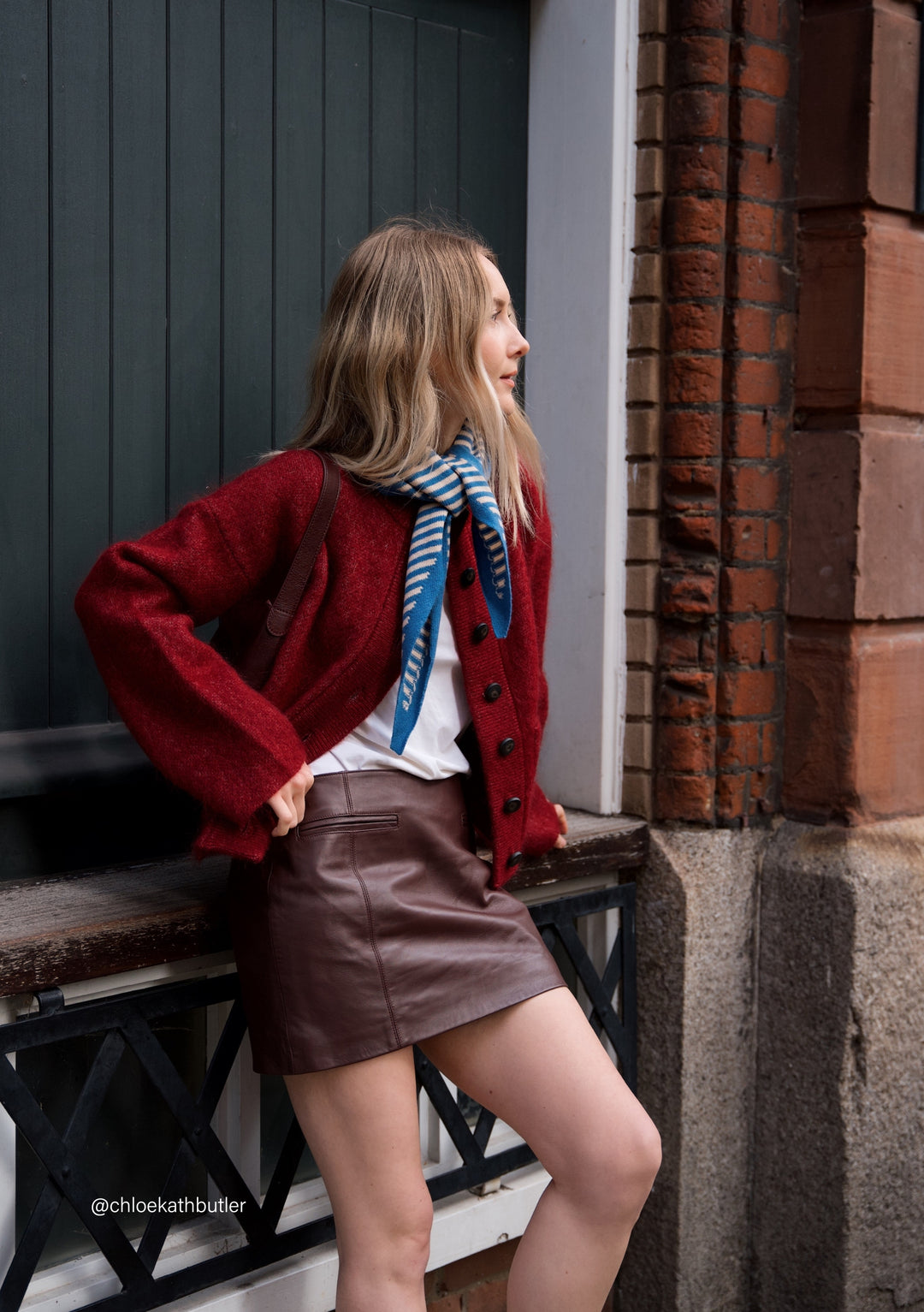 TBCo Merino Triangle Scarf in blue and cream stripes styled with red cardigan, showcasing versatile winter layering against brick wall