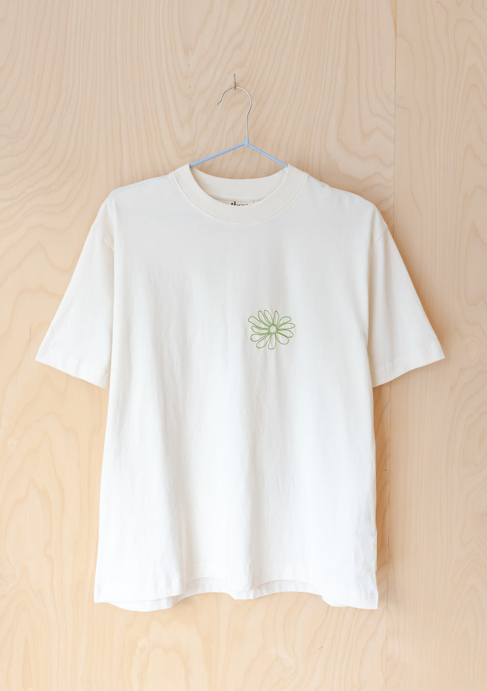 TBCo Cotton T-Shirt in Ecru Flora with embroidered green daisy and floral checkerboard back print, hanging on wooden wall