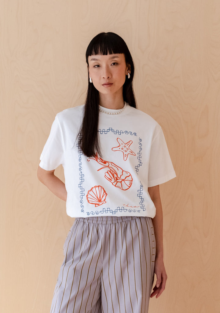 White cotton t-shirt from TBCo featuring red shrimp and sea life illustrations with blue border design, styled against beige backdrop