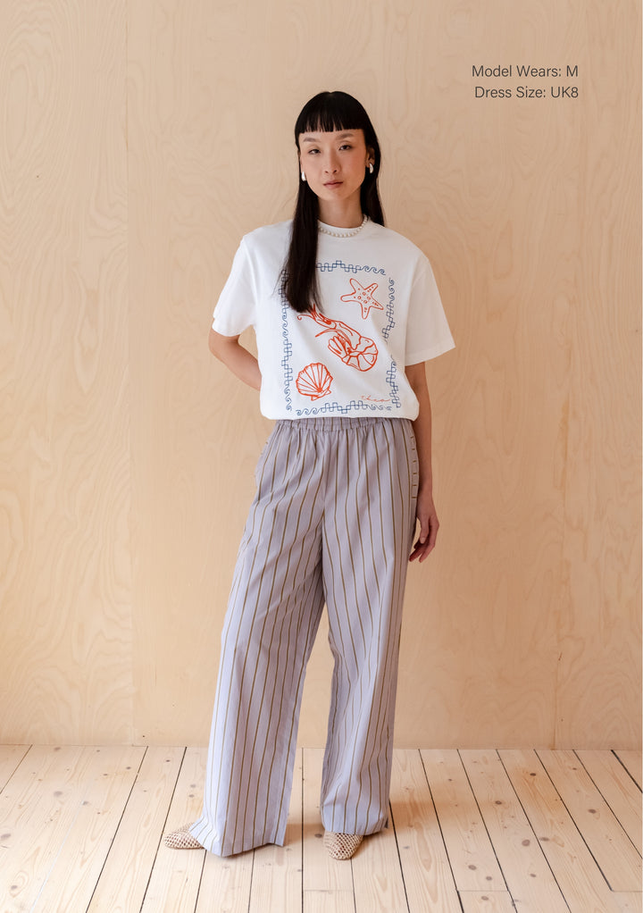 White cotton TBCo t-shirt with red shrimp and shell illustration in blue border, paired with grey striped trousers