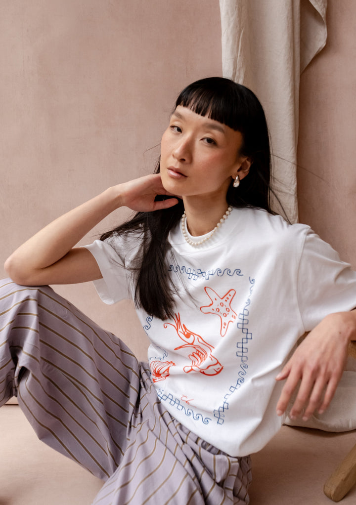 A cotton t-shirt by TBCo featuring a whimsical red shrimp design with blue waves, styled with pearl necklace against blush backdrop.