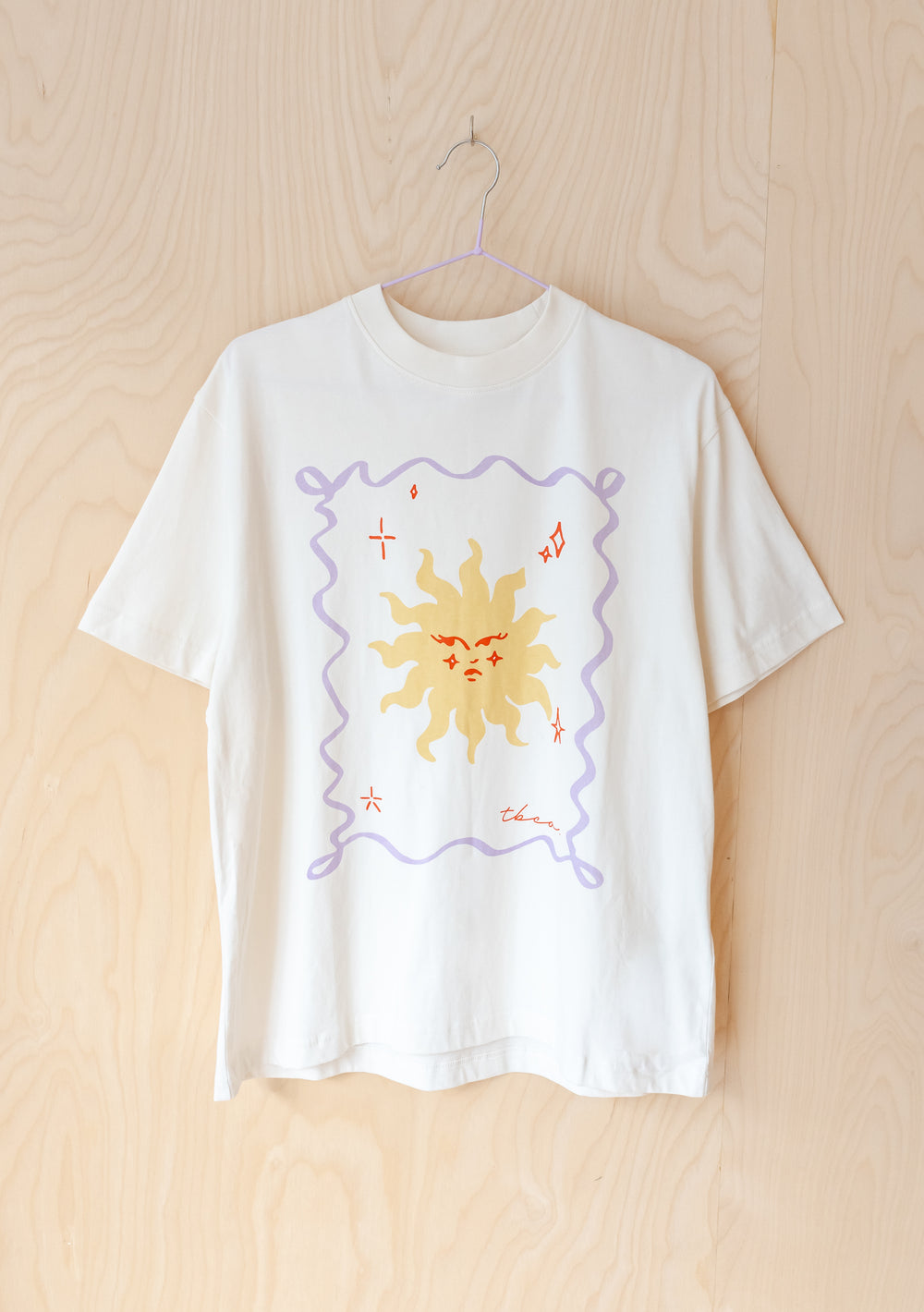 TBCo Cotton T-Shirt in Ecru Sun featuring yellow sun illustration with lilac border on cream cotton, displayed on hanger