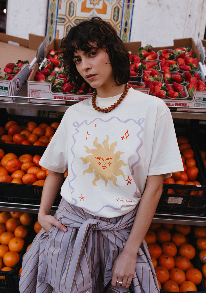 TBCo Cotton T-Shirt in Ecru Sun with illustrated yellow sun design and lilac border, shown against fresh produce display