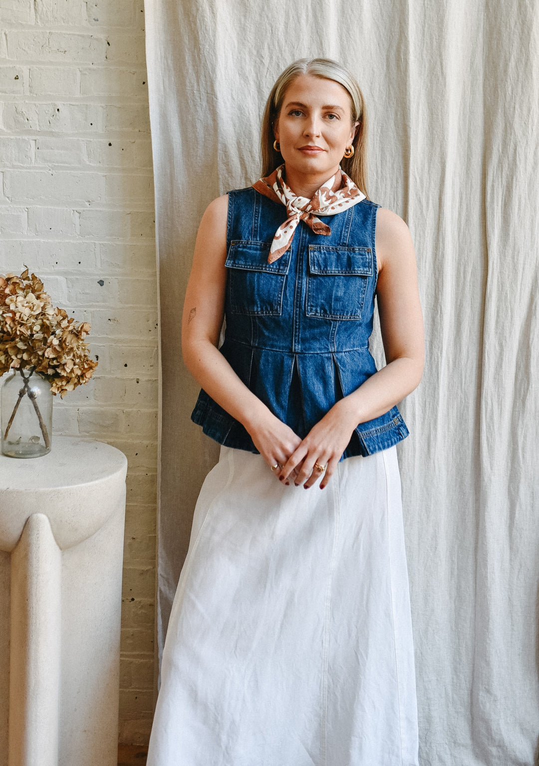Woman wearing The Artist Series TBCo x Frankie Penwill Cotton T-Shirt, styled with a patterned scarf and skirt, stands beside a table with dried hydrangeas.