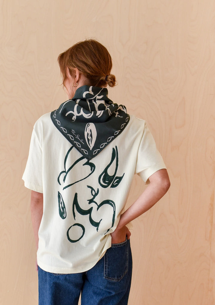 Person in The Artist Series TBCo x Simone Brewster Cotton T-Shirt with abstract brushstroke design, wearing a dark bandana, standing against a wooden wall.