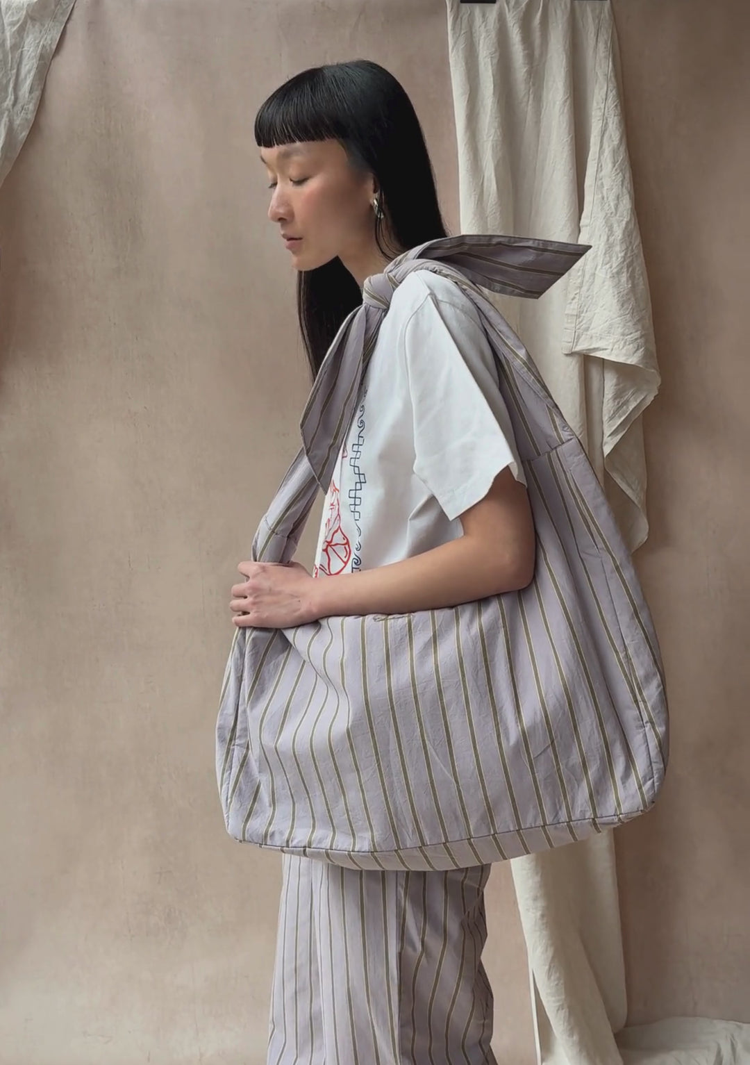 A model poses against a neutral ply backdrop, wearing TBCo matching lilac stripe trousers and oversized shoulder bag, with a TBCo white t-shirt.