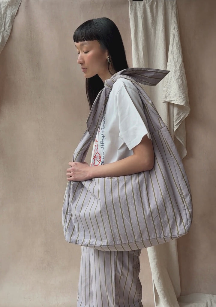 A model poses against a neutral ply backdrop, wearing TBCo matching lilac stripe trousers and oversized shoulder bag, with a TBCo white t-shirt.