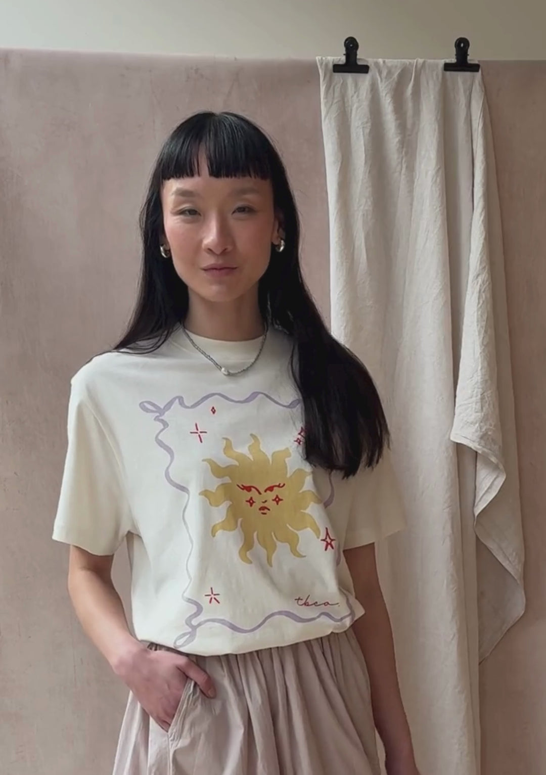 A model poses against a neutral ply backdrop, wearing a TBCo cream t-shirt with a yellow sun and lilac border motif.
