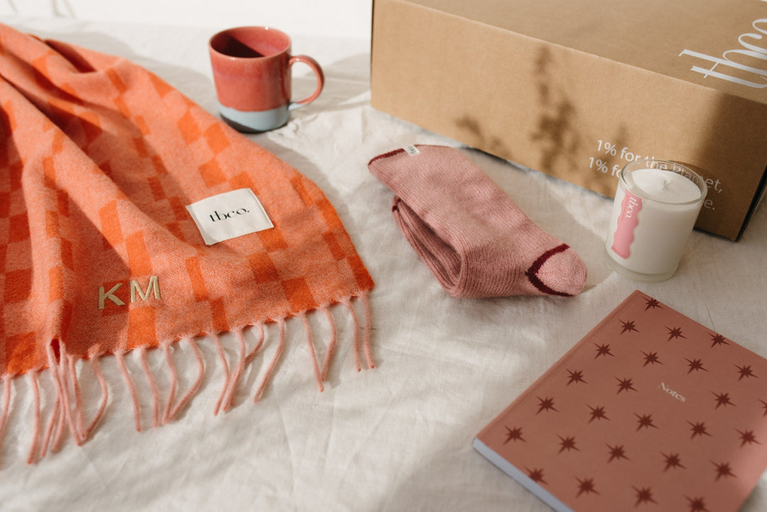 A TBCo gift box with contents including a TBCo lambswool orange blanket, mug, socks, candle and notebook.