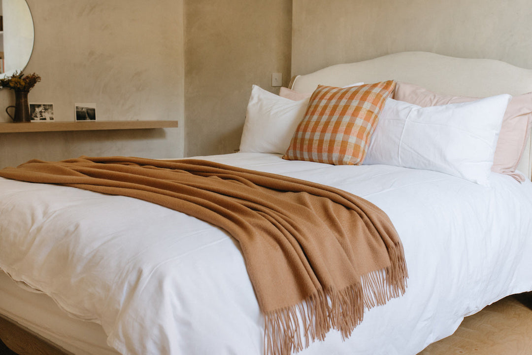 A TBCo camel cashmere blanket draped across a neutral bed.