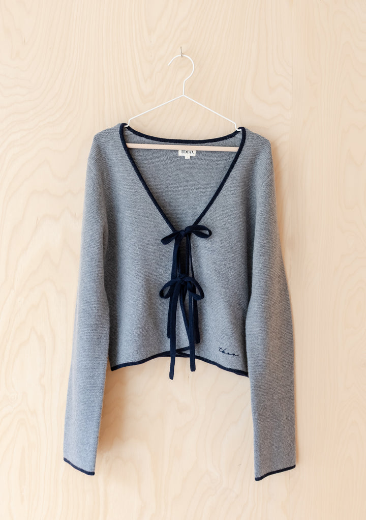 TBCo Merino Wool Tie Cardigan in Grey: Light-grey knit cardigan with navy trim, V-neck, and tie closures, featuring TBCo logo. Crafted from 100% merino wool for versatile layering.