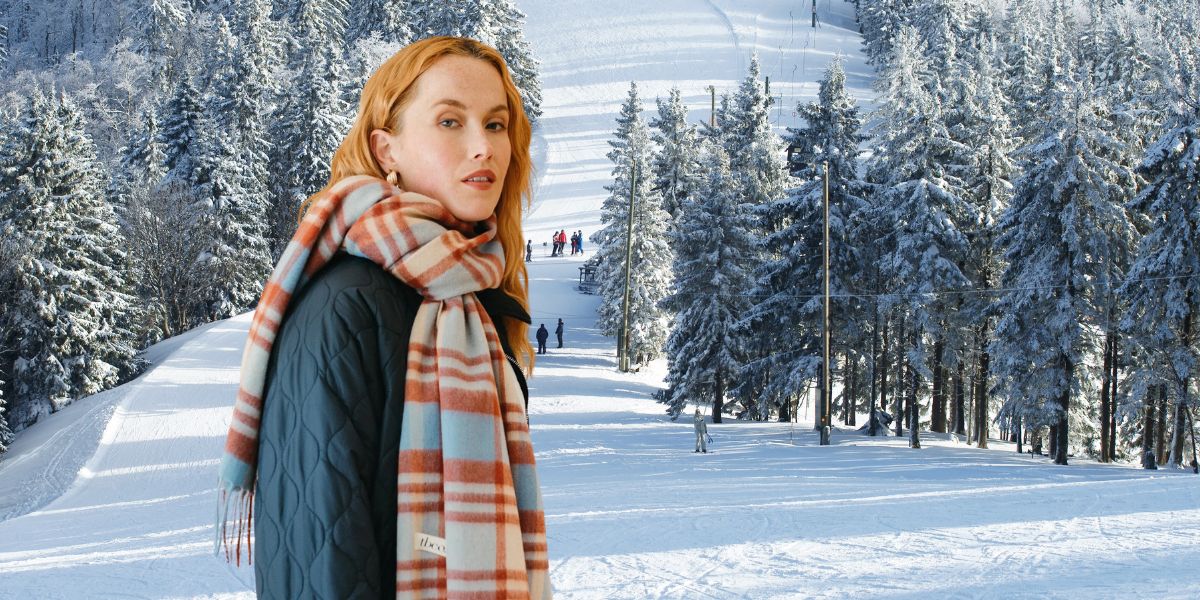 TBCo Lambswool oversized lilac multi check scarf wrapped around the neck, styled with a grey coat in front of a snowy  apres ski backdrop. A soft and elegant  accessory for timeless layering