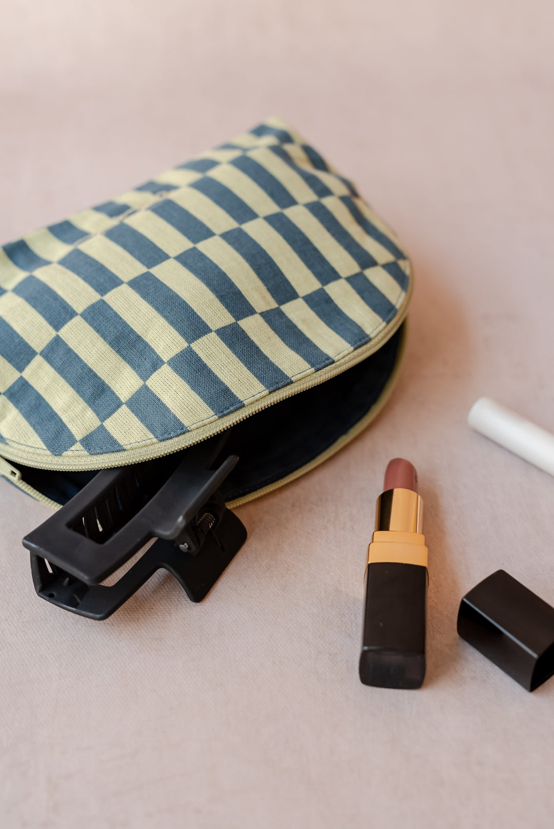A TBCo make-up pouch in a green checkerboard pattern sits on a neutral ply backdrop with a hair clip, lipstick and makeup brush all sprawling out of it.