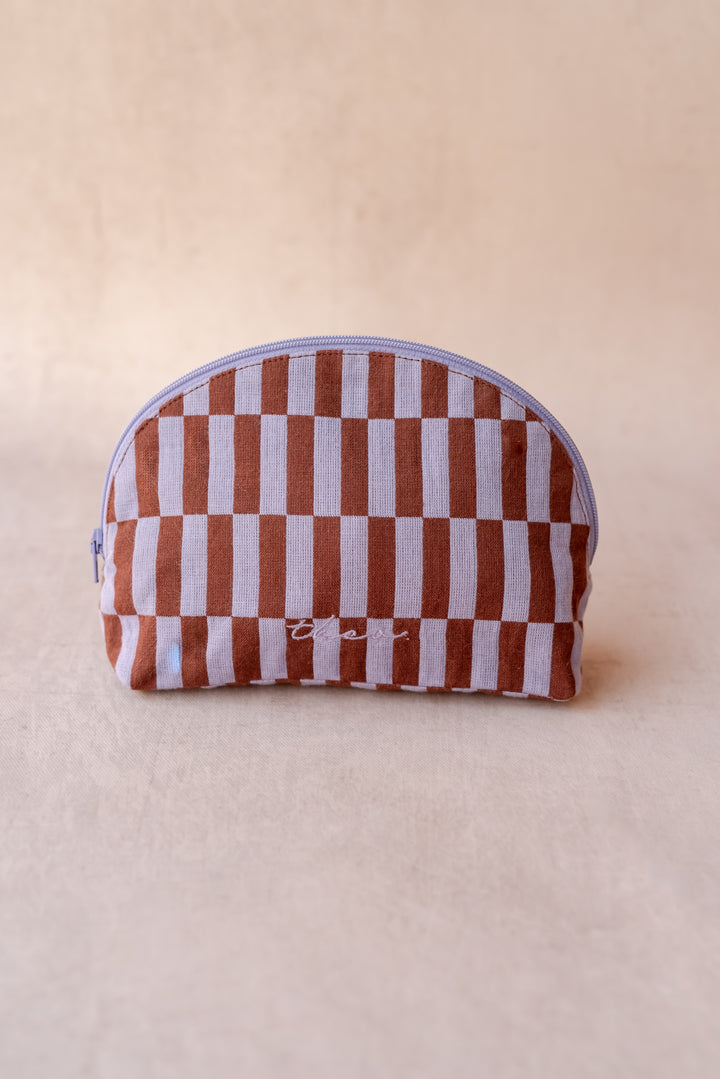 A TBCo make-up pouch in a lilac and rust checkerboard pattern sits against a neutral ply backdrop