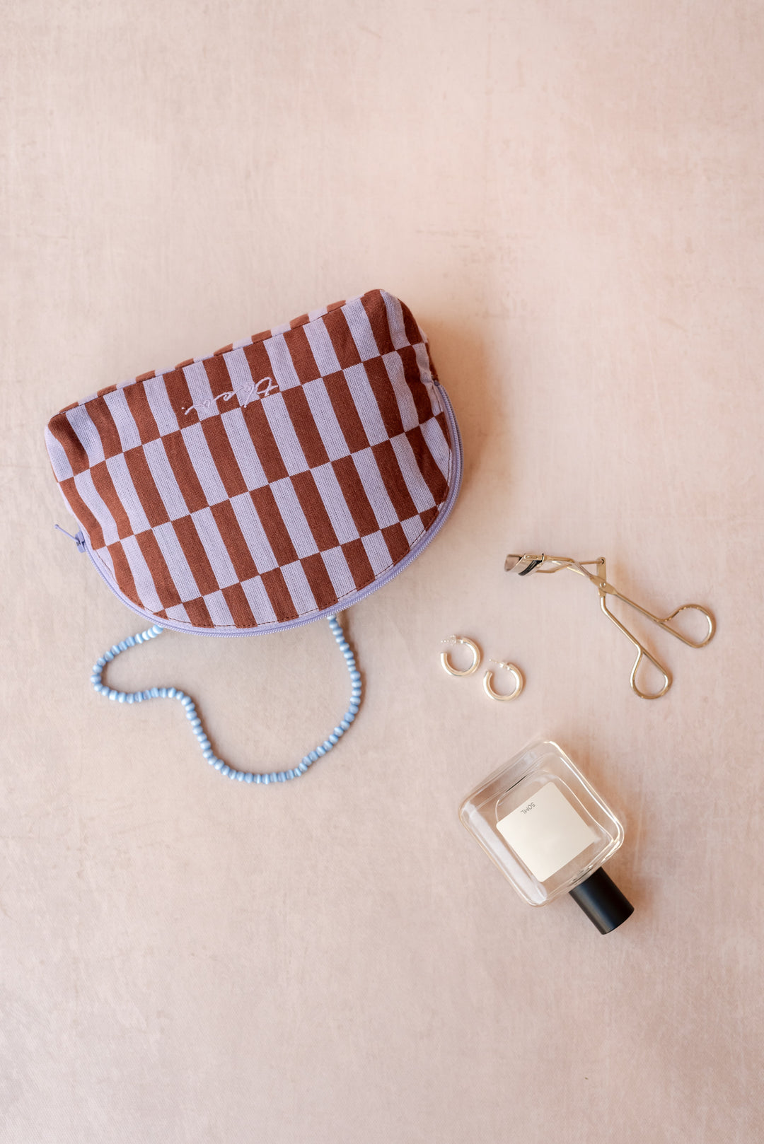 A TBCo make-up pouch in a lilac and rust checkerboard pattern sits on a neutral ply backdrop with a blue necklace, perfume bottle, eyelash curlers and earrings all sprawling out of it.