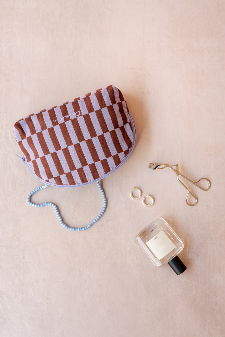 A TBCo make-up pouch in a lilac and rust checkerboard pattern sits on a neutral ply backdrop with a blue necklace, perfume bottle, eyelash curlers and earrings all sprawling out of it.
