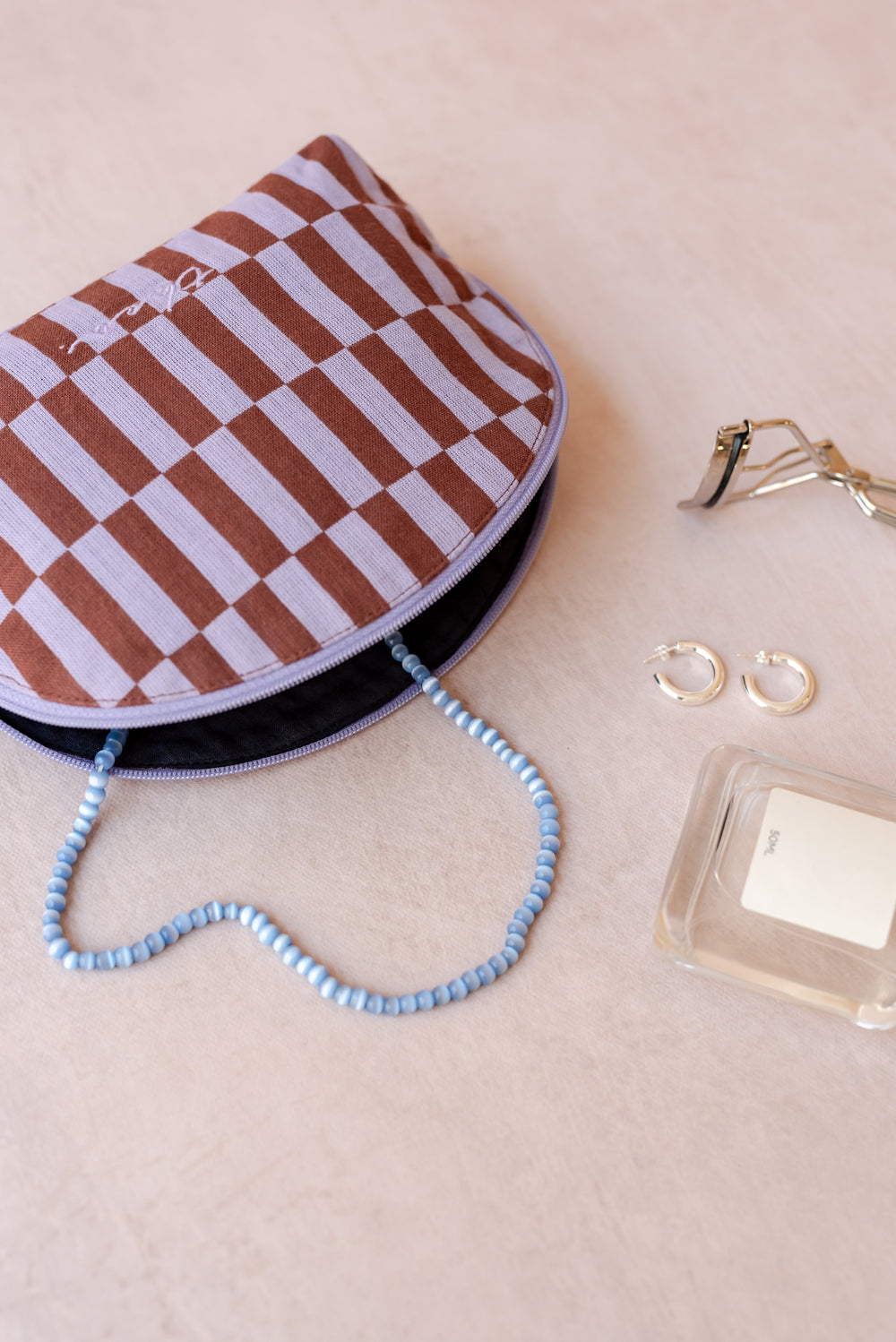 A TBCo make-up pouch in a lilac and rust checkerboard pattern sits on a neutral ply backdrop with a blue necklace, perfume bottle, eyelash curlers and earrings all sprawling out of it.