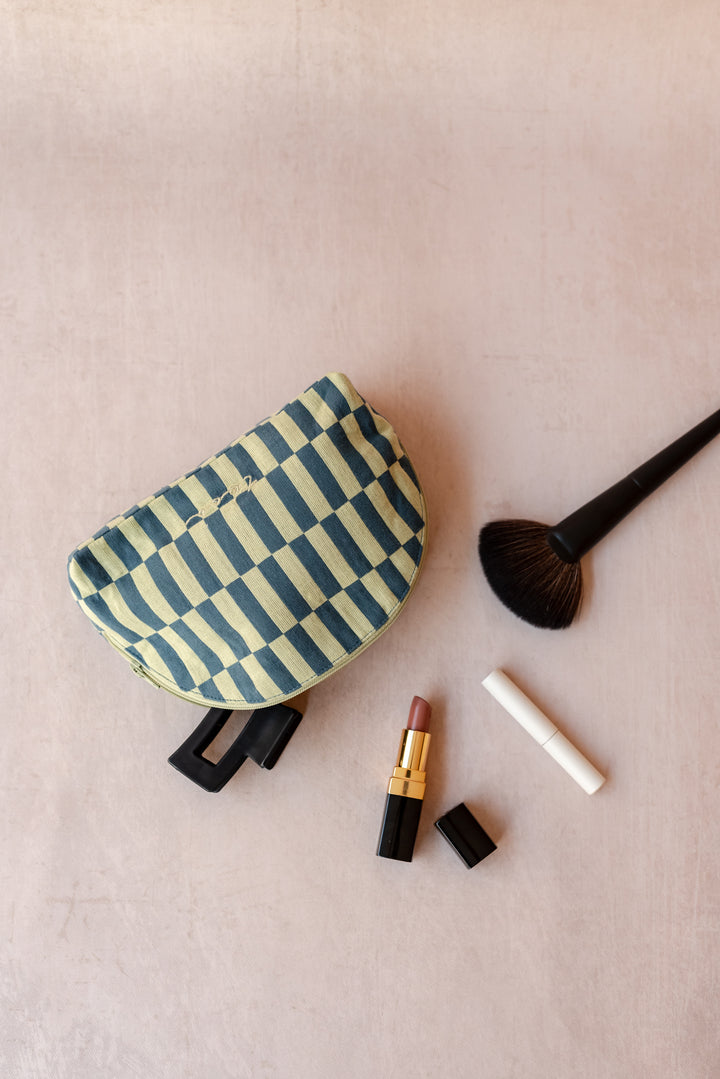 A TBCo make-up pouch in a green checkerboard pattern sits on a neutral ply backdrop with a hair clip, lipstick and makeup brush all sprawling out of it.
