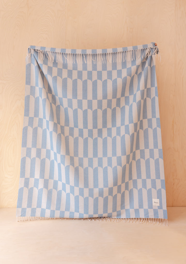 TBCo Lambswool Blanket in Blue Geo hanging against pale wood, featuring soft fringes and a light blue geometric pattern