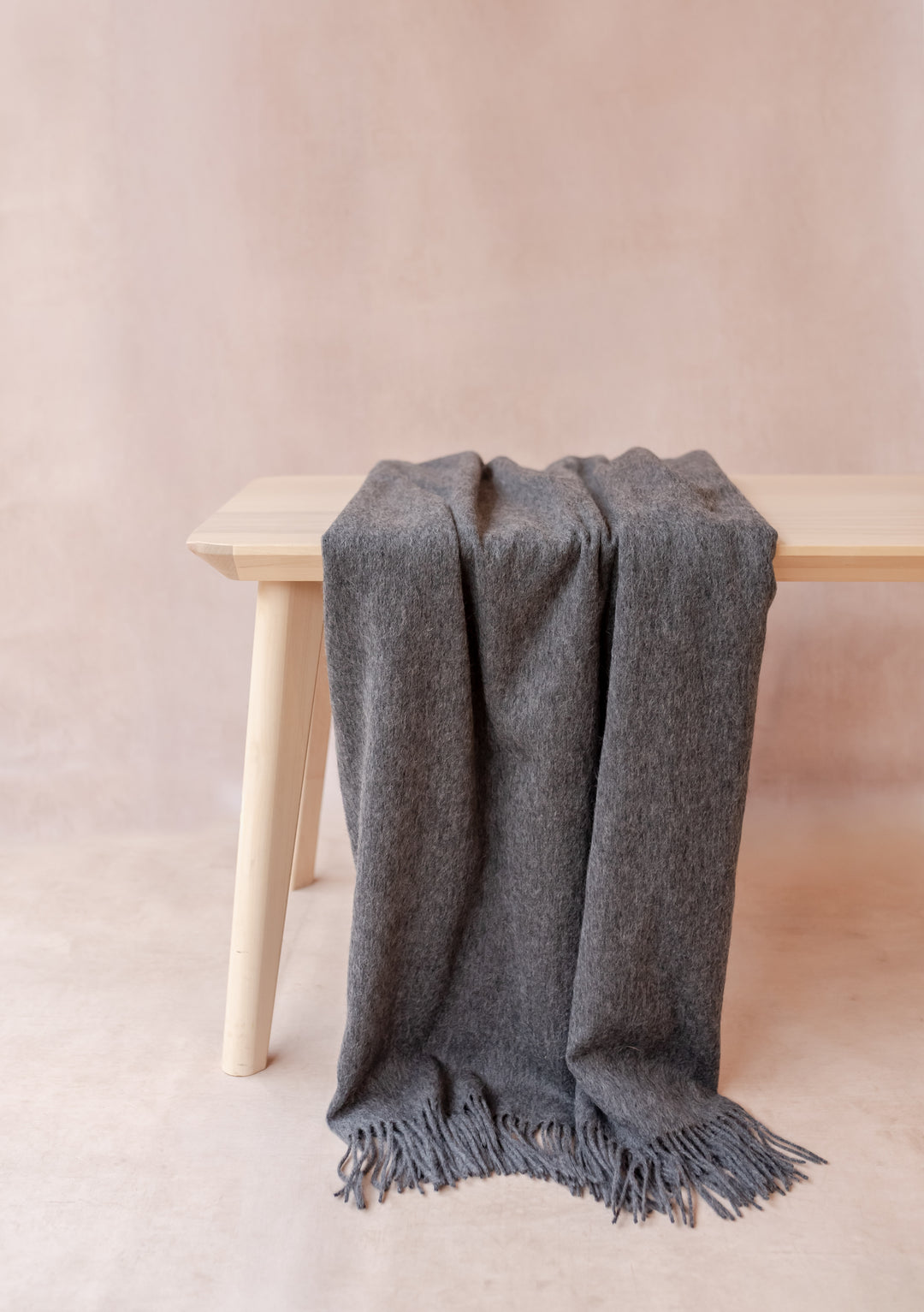 Cashmere Small Blanket in Charcoal Melange by TBCo draped over a light wooden bench with soft fringe detailing