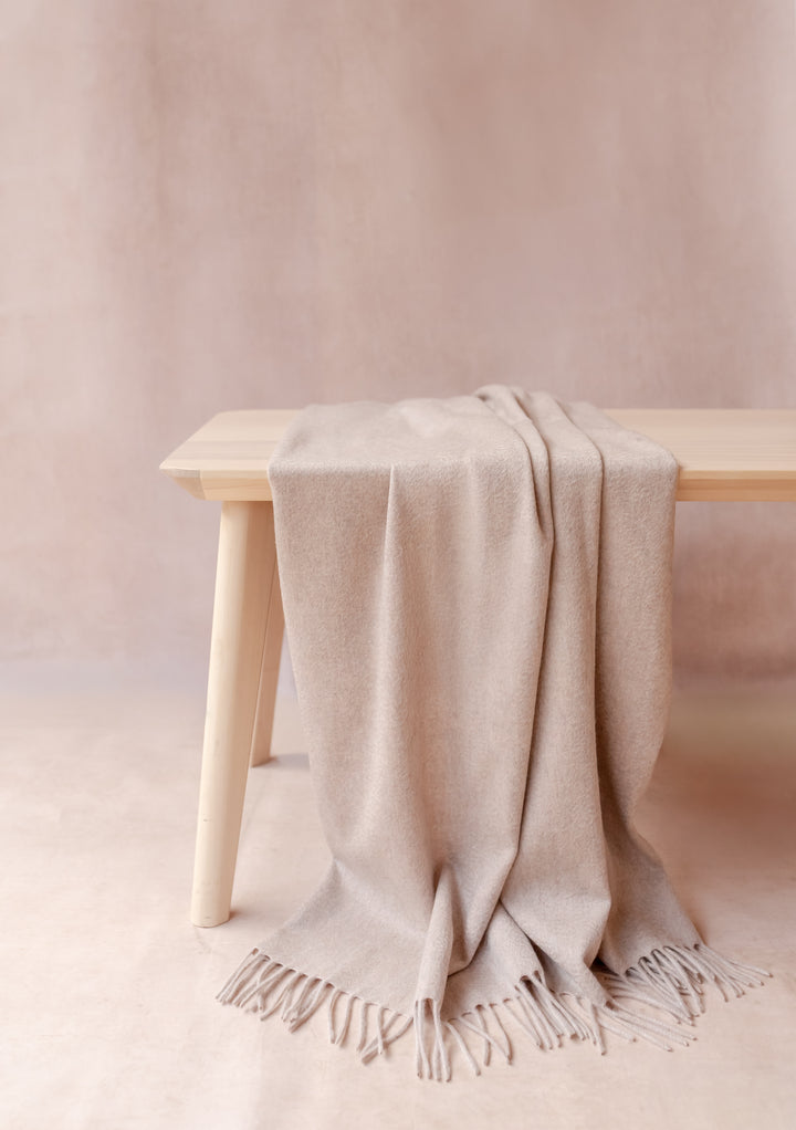 TBCo Cashmere Small Blanket in Oatmeal Melange draped over a wooden bench showcasing its soft texture and fringe edges