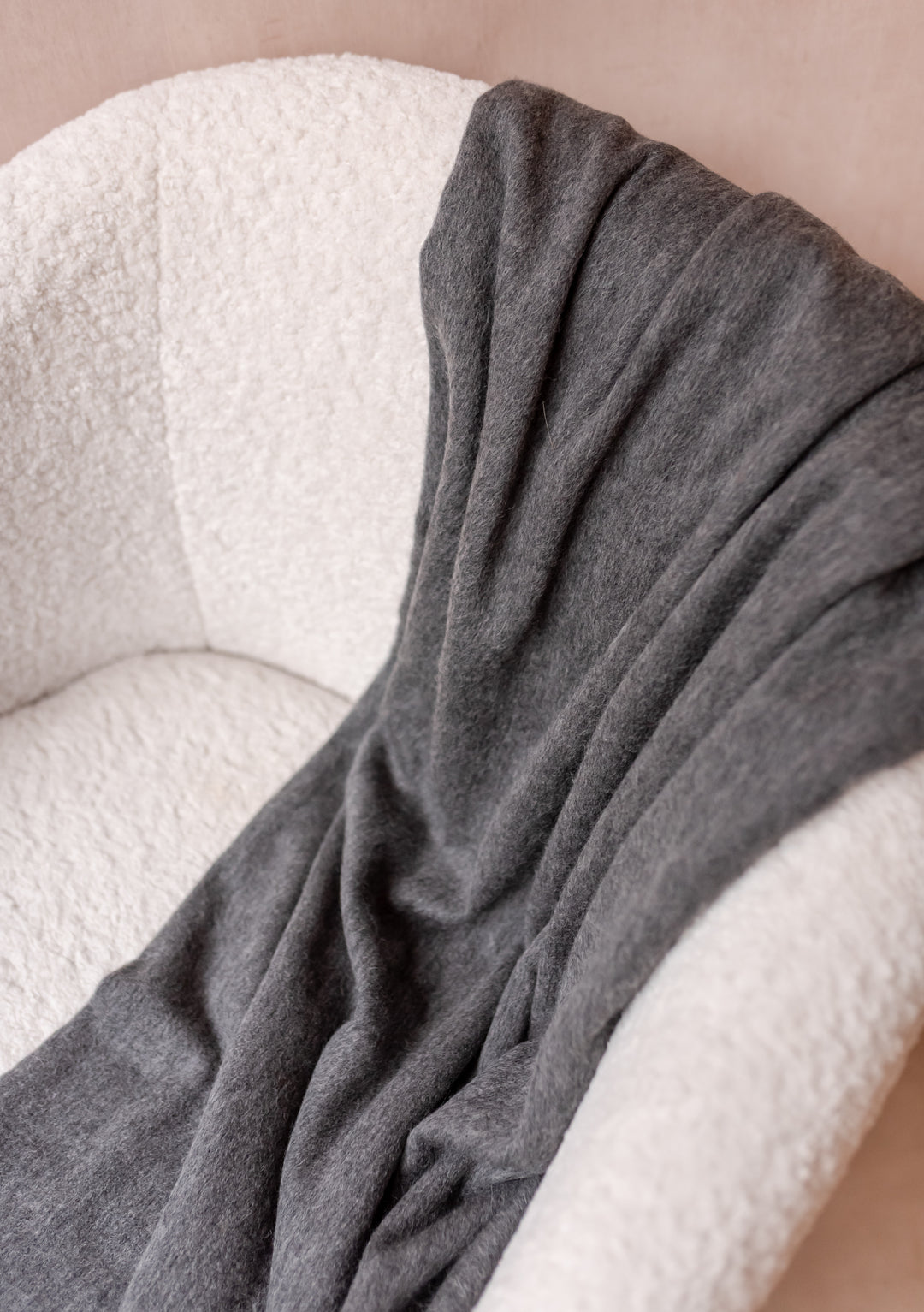 TBCo Cashmere Blanket in Charcoal Melange draped over a white textured armchair showcasing soft, luxurious fabric with fringe edges.