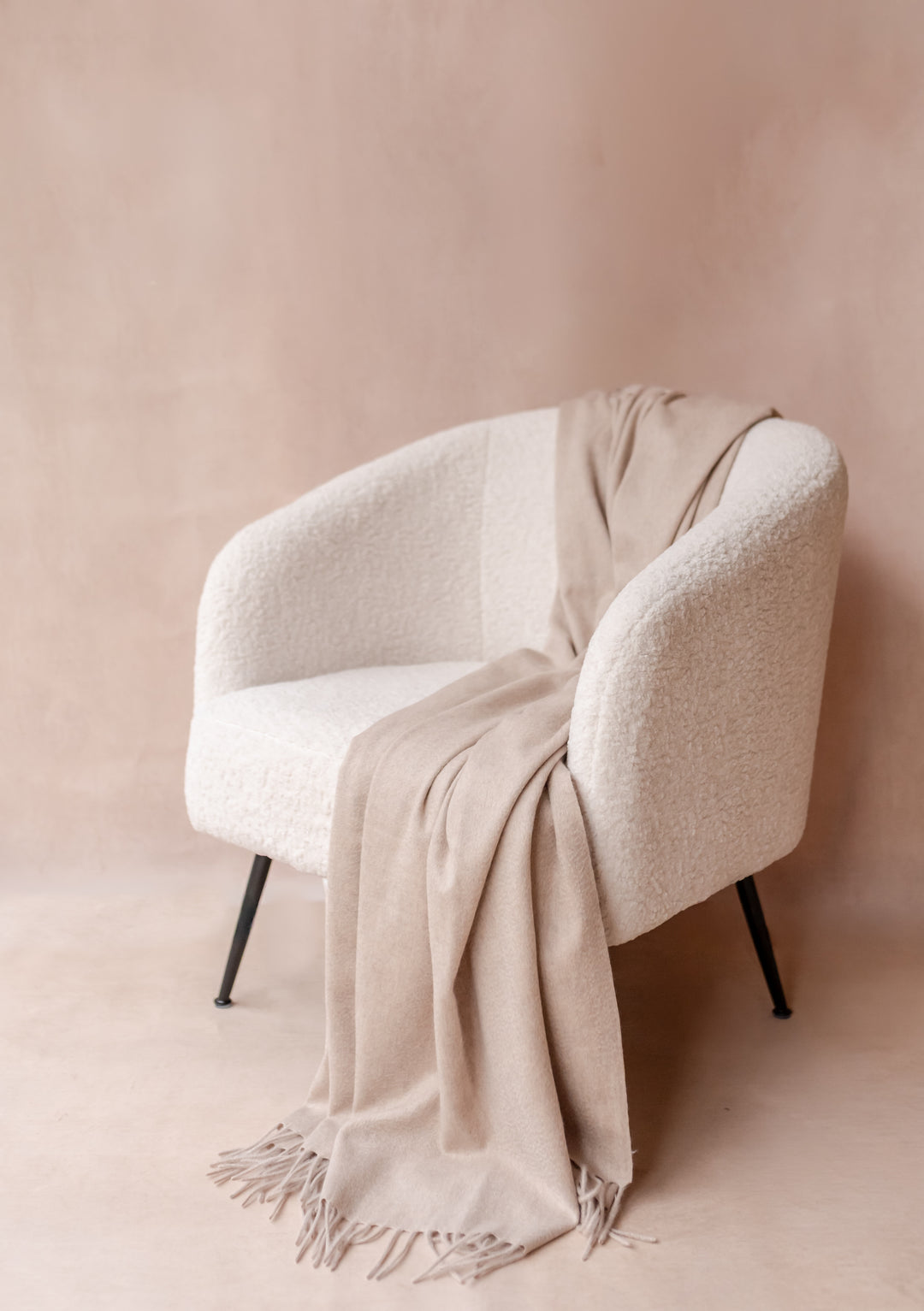 Cashmere Blanket in Oatmeal Melange by TBCo elegantly draped over a textured white armchair with soft fringe detailing