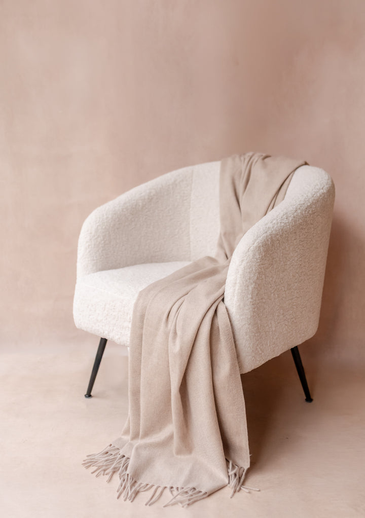 Cashmere Blanket in Oatmeal Melange by TBCo elegantly draped over a textured white armchair with soft fringe detailing