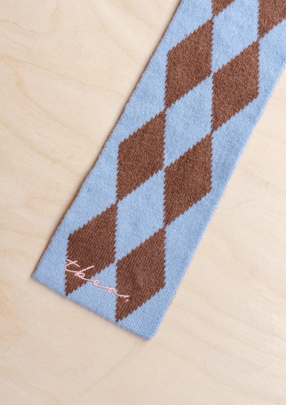 Close-up of TBCo Merino Wool Skinny Scarf in Brown Harlequin showing blue and brown diamond pattern on light wood surface