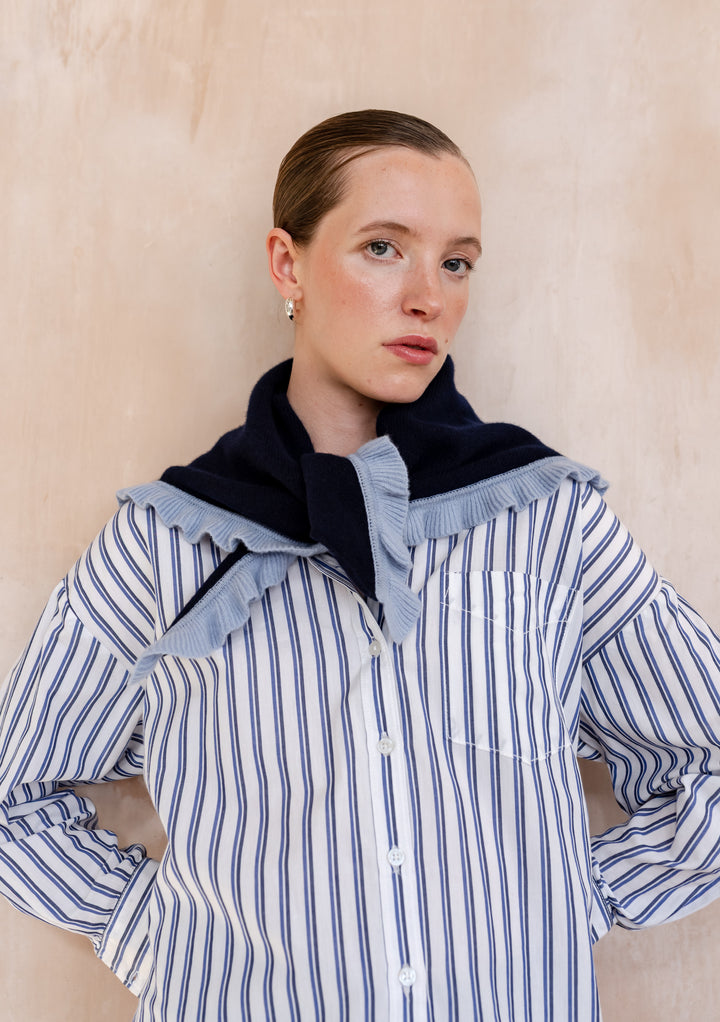 Woman models TBCo Merino Wool Triangle Scarf in Navy Ruffle, draped over shoulders, showcasing its light-blue ruffle trim and soft merino wool texture.