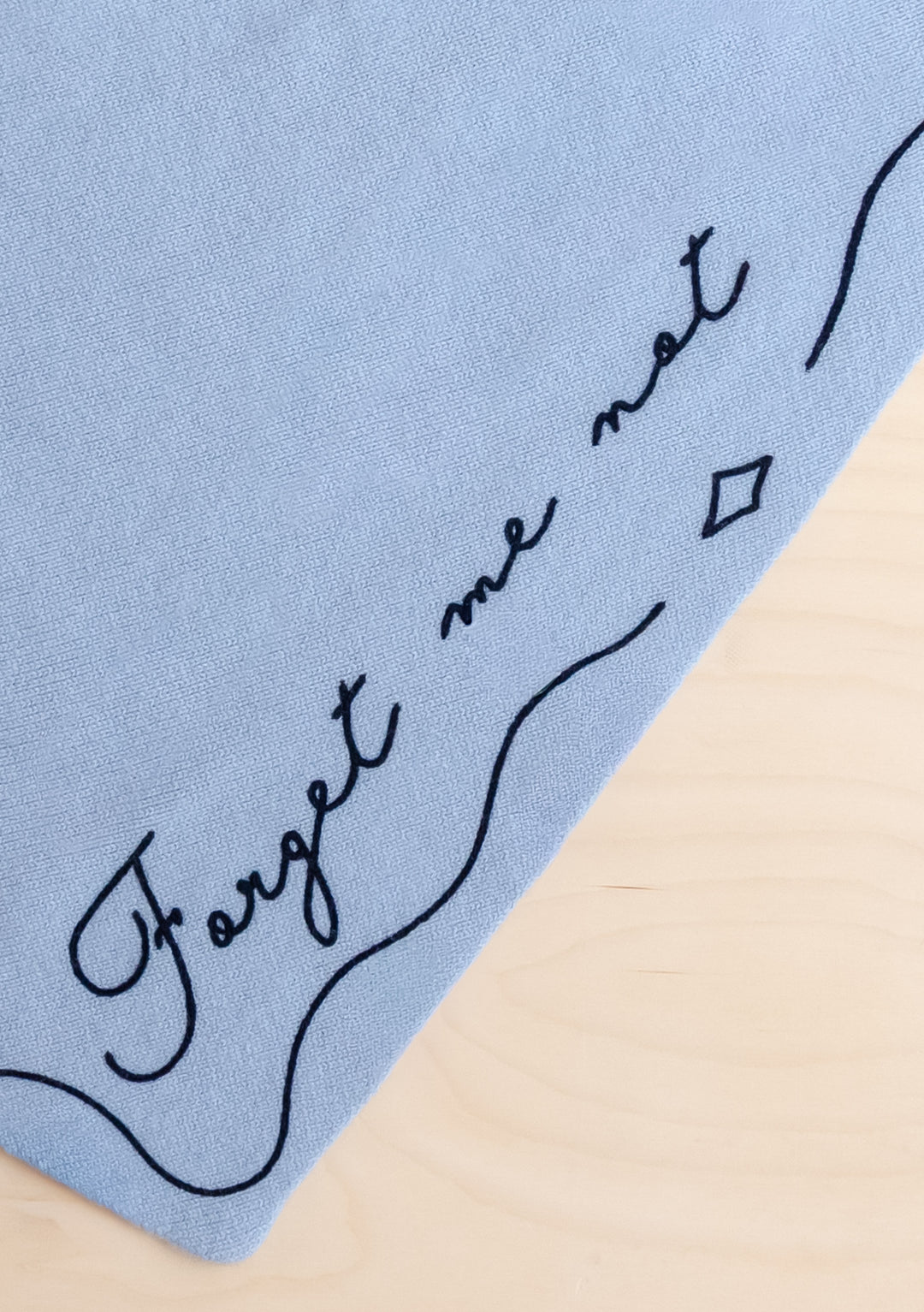 Close-up of TBCo Merino Wool Triangle Scarf in Forget Me Not showing pale blue fabric with embroidered text detail