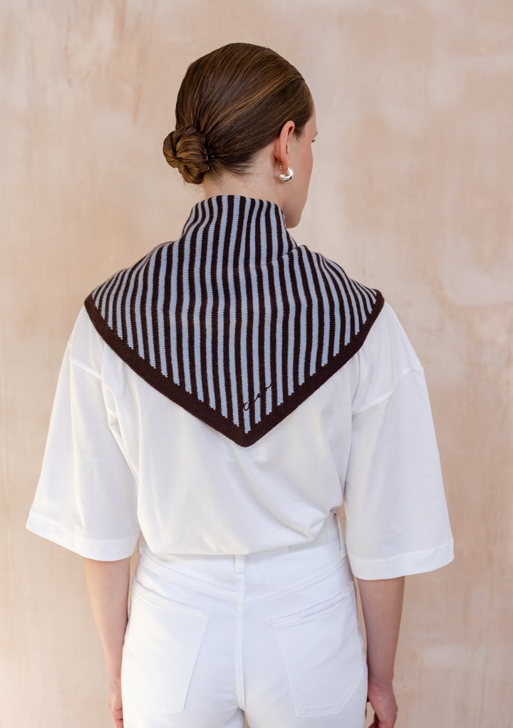 Person wearing TBCo Merino Wool Triangle Scarf in Blue & Brown Stripe, draped over shoulders against a light-wood background, highlighting its stylish, versatile design.