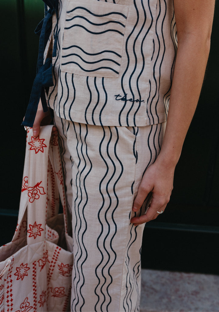 A model wears a TBCo two piece trouser set in white and blue wave pattern, holding a white and orange TBCo tote bag and standing in front of a green door on a Lisbon street.