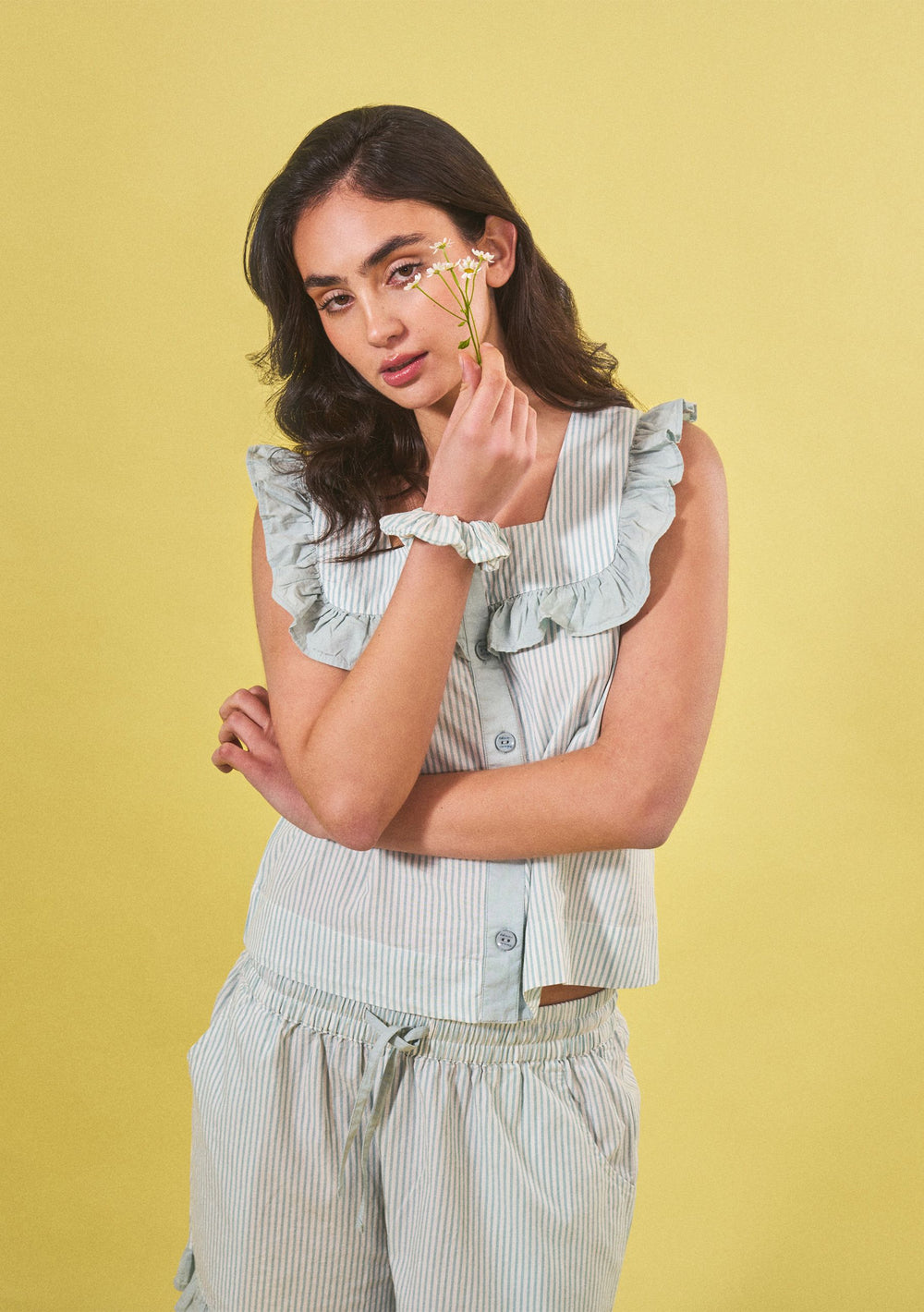 Model wearing Cotton Pyjamas in Blue Stripe Ruffle by TBCo in portrait, holding white flowers against yellow background