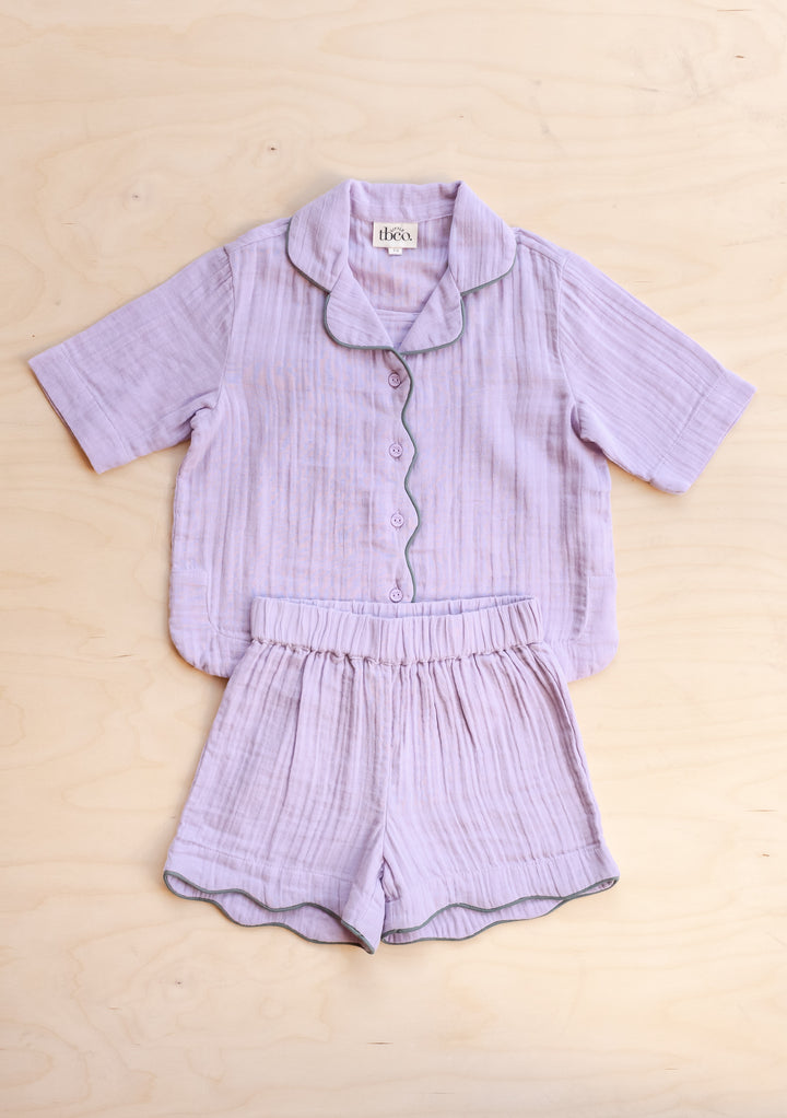 TBCo Cotton Kids Pyjamas in Lilac with short sleeves, scalloped trim, button-up top, and elasticated shorts on pale wooden surface