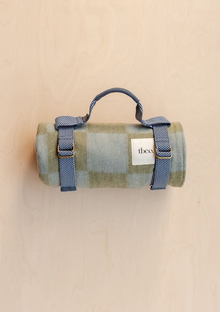 Rolled TBCo Recycled Wool Picnic Blanket in Green & Blue Checkerboard with blue buckle straps on light wood background