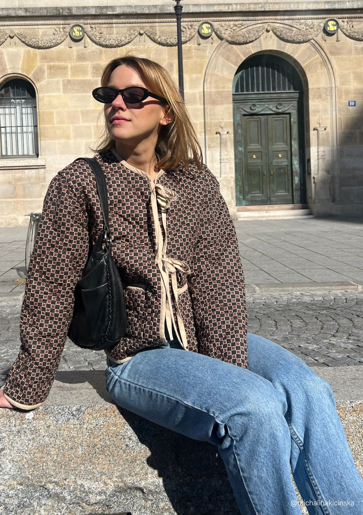 A model poses on a wall in Paris wearing a TBCo brown cotton quilted jacket, blue jeans and black shoulder bag.