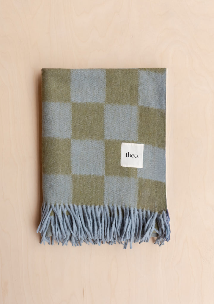 Portrait shot of folded TBCo Recycled Wool Blanket in Blue & Green Checkerboard with fringed edges on wooden surface.