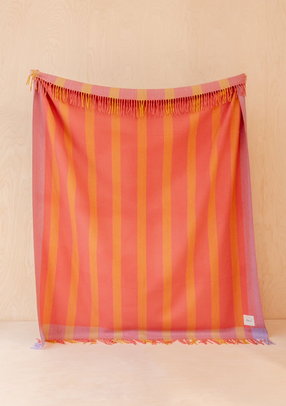 TBCo Recycled Wool Blanket in Orange Frame Stripe hanging with delicate fringed edges against pale wooden wall