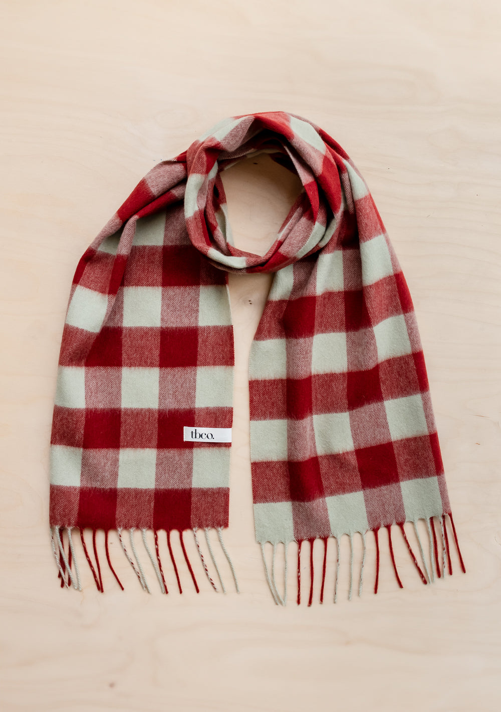 TBCo Lambswool Scarf in Red Gingham featuring sage green and red check pattern with fringed ends, laid flat on beige surface