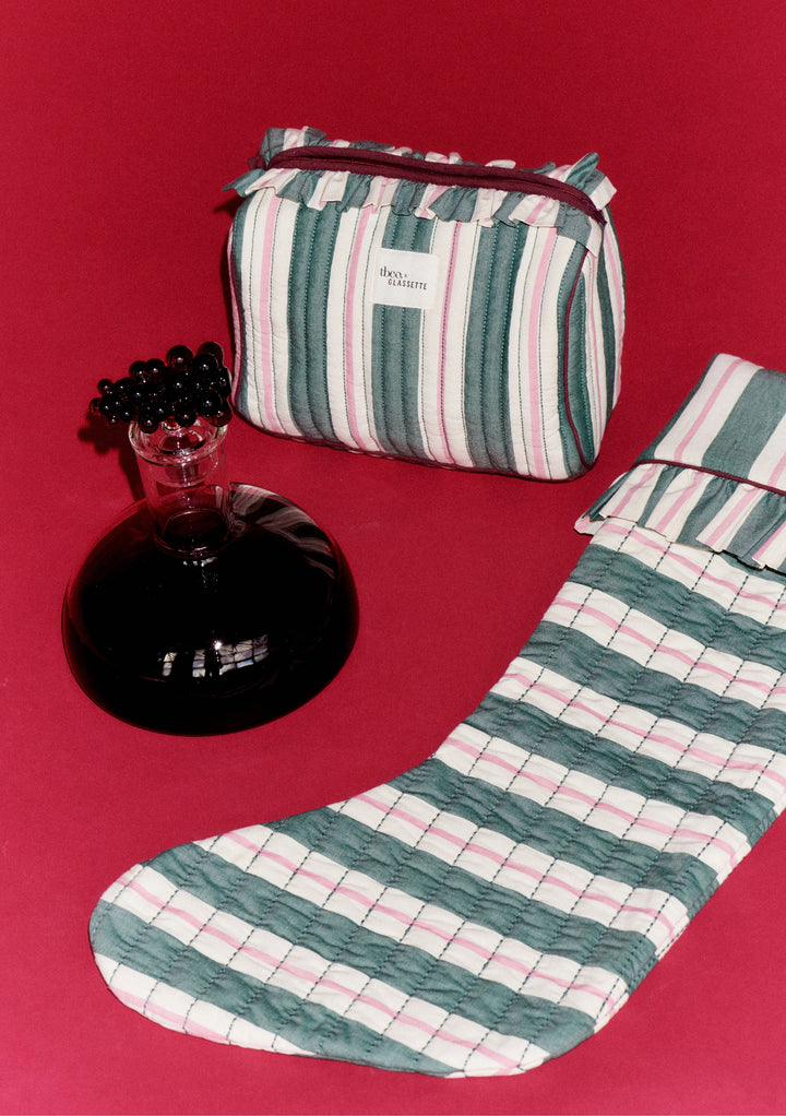 TBCo x Glassette Quilted Cotton Christmas Stocking in Green Stripe with ruffle trim, paired with matching pouch and dark glass decanter on a red background.
