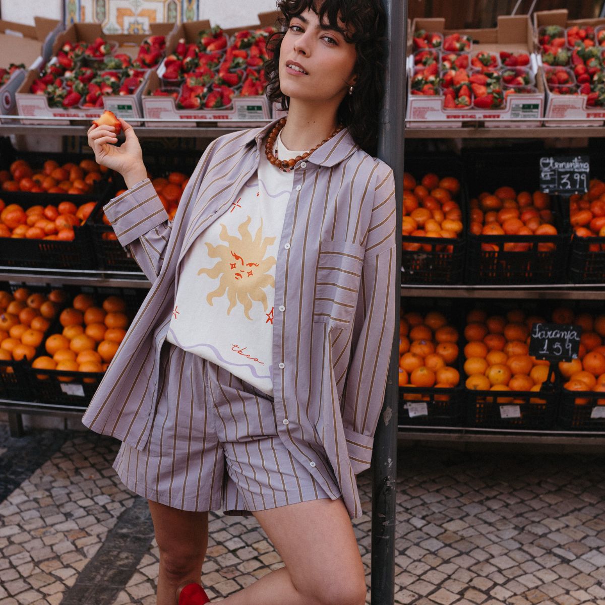 Woman in a TBCo Cotton Lilac Stripe shirt and shorts co-ord set with sun-printed white cotton t-shirt, posing confidently at a vibrant fruit market with crates of oranges and strawberries in the background.